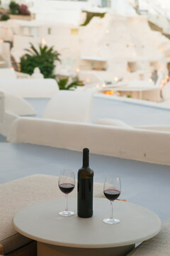 A bottle of red wine and two glasses rest on a terrace at dusk in Santorini, Greece, evoking Mediterranean elegance and quiet evening relaxation.