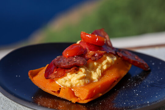 A Mediterranean breakfast of scrambled eggs, tomatoes, and cured meats served on a sweet potato in Santorini, Greece, glowing in warm coastal sunlight.