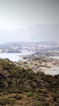 The river moves through a rocky area with scattered trees. The sun shines on the water and land. Mountains are in the background. It is daytime and the scene shows natural beauty.