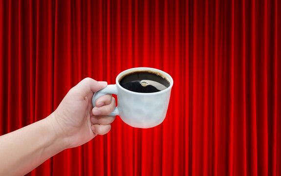 A white ceramic coffee cup is in a man's hand, against a background of a red curtain.