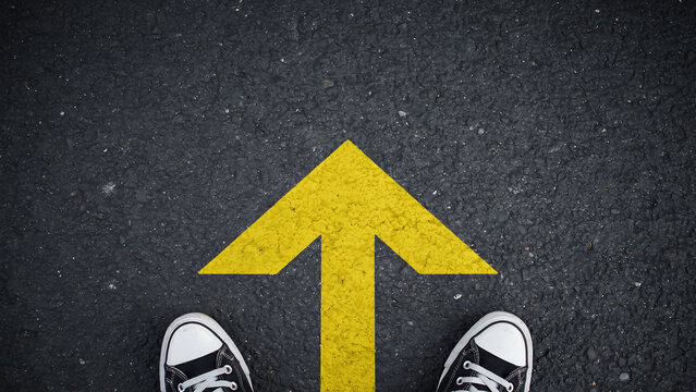 Casual shoes standing on asphalt ground next to a bright yellow direction arrow painted on the surface with direction navigation guidance lifestyle movement opportunity adventure decision