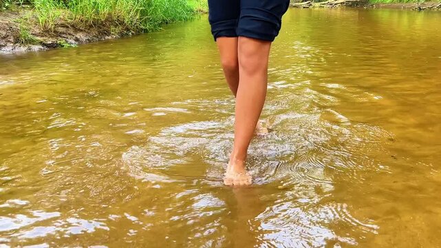 Low angle shot of a person walking barefoot through a shallow, flowing river stream in a natural forest setting. Suitable for outdoor, travel, and wellness projects.