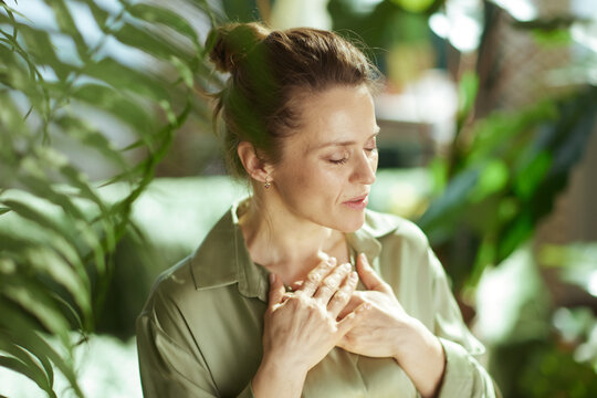 Embodying tranquility, a woman stands with eyes closed and hands over her heart, surrounded by lush greenery and soft sunlight for a moment of emotional balance.