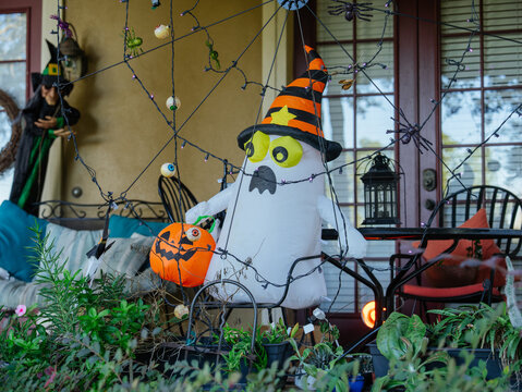A Halloween front porch features a large inflatable ghost wearing a witch hat surrounded by spider web decorations and glowing lights. Pumpkins, spiders, eyeballs, and seasonal decorations create a