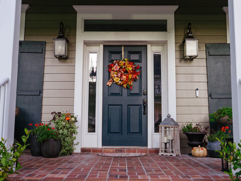 A colorful autumn wreath hangs on the front door of a modern residential home. Seasonal flowers, pumpkins, and decorative plants create a warm fall entrance.