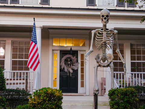 A towering Halloween skeleton stands beside the entrance of a residential home with seasonal decorations. The spooky outdoor display creates a festive Halloween scene in a suburban neighborhood.