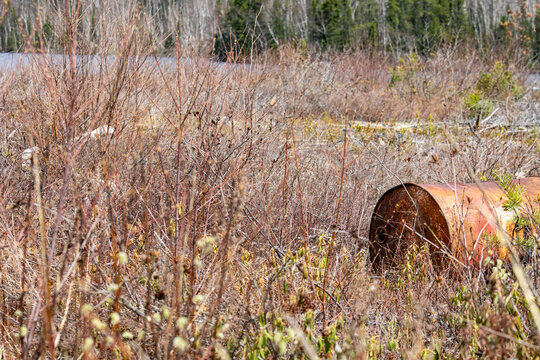 Parents, Quebec, 20 May 2026 : A rusty metal barrel lies abandoned amidst dry brush and sparse vegetation near a body of water.