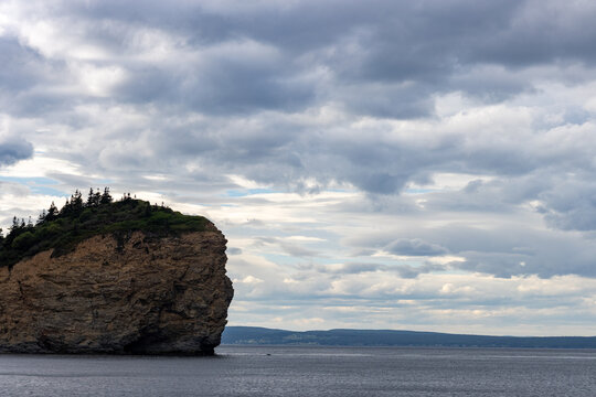 Gaspesie, Quebec, 15 July 2026 : A large rocky cliff covered with trees rises from the water under a cloudy sky