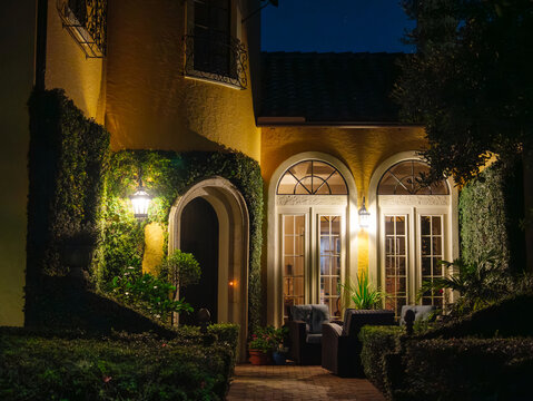 A beautifully illuminated courtyard entrance features arched doorways, landscaped hedges, and cozy outdoor seating. Warm lighting highlights the timeless architecture of the luxury residence after