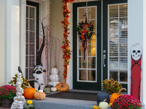 A front door is surrounded by pumpkins, a Halloween wreath, autumn leaves, and illuminated seasonal decorations. A festive display welcomes visitors with classic fall holiday decorations.
