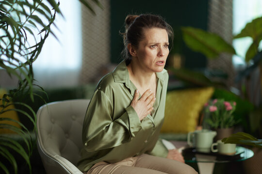 A woman in a sage green blouse sits in a sunlit living room, holding her hand to her chest while experiencing shortness of breath or anxiety in a cozy, plant-filled home setting.