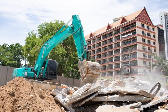 Excavator with bucket clearing concrete debris at demolition site near apartment building