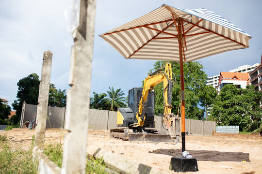 Yellow Excavator With Hydraulic Breaker Attachment Parked At Sandy Construction Site Near Sun Umbrella Under Blue Sky