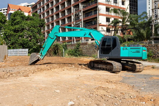 Teal excavator with bucket digging soil at construction site near apartment building