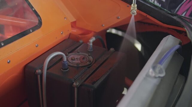 Action shot of a water spray nozzle rinsing down the radiator and fuel cell compartment inside the orange-painted engine bay of a race car.