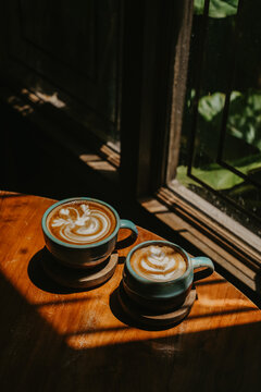 Moody aesthetic coffee photography showcasing two latte cups on wooden coasters, illuminated by strong window light and sharp shadows