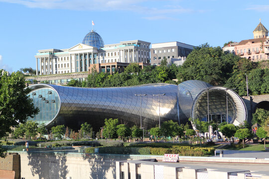 Modern metallic tube-shaped concert hall and exhibition center in Rike Park Tbilisi Georgia with Glass and metal dome Presidential Palace on hill background blue sky urban architecture landscape