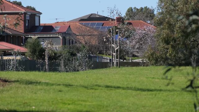 A grassy public lawn toward residential homes in Melton, Melbourne, Australia.&nbsp;Houses stand behind timber boundary fences, framed by young native trees and public green space