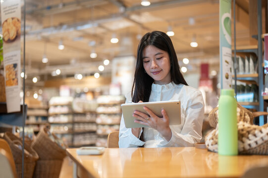 Young asian woman writing in tablet while sitting with a bag and tumbler at table in a shopping mall