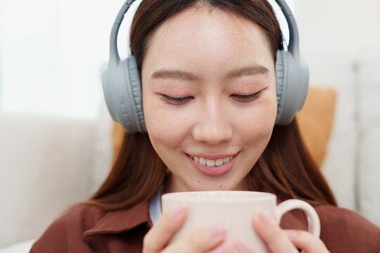 Close up of young, beautiful Japanese woman in brown shirt listening to music with headphones, holding and looking at coffee or tea mug with both hands, smelling with gentle smile in cozy living room 