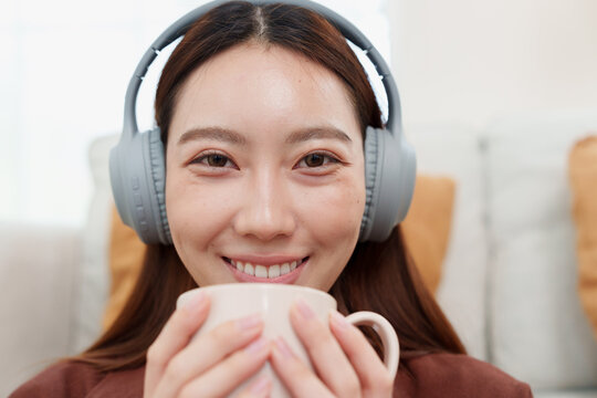 Close up of young, beautiful Japanese woman in brown shirt with headphones looking at camera, holding warm coffee or tea mug with both hands, smelling with gentle smile in cozy living room at home