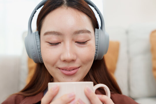 Close up of young, beautiful Japanese woman in brown shirt with headphones holding warm coffee or tea mug with both hands, smelling with gentle smile in cozy living room at home
