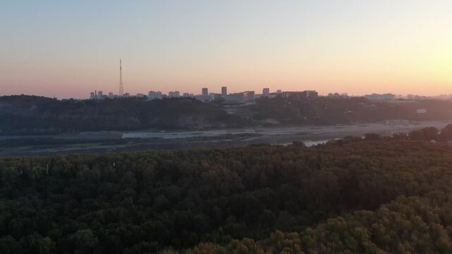 Aerial view of the Belaya River at sunrise, featuring dense green forests along the banks and the urban skyline in the background in Ufa, Russia.
