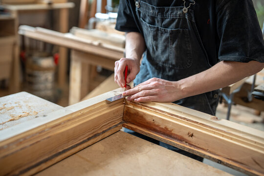 Carpenter working at workbench in restoration workshop on final fitting of antique window frame part, professional woodworker at table using ruler and pencil checking alignment of corners and fixation