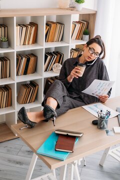 Digital work. Corporate routine. Business woman drinking coffee talking on smartphone at desk with notebook reviewing document at professional workplace.