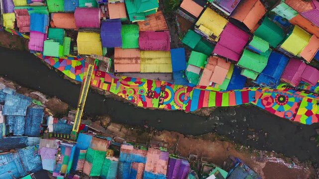 Colorful rooftops dominate this vibrant slum settlement. Winding paths cut through densely packed structures. Muddy river runs alongside the crowded dwellings. Buildings show varied hues
