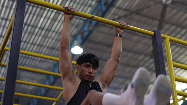 Muscular Asian man performing hanging leg raises. Athletic young Asian man doing hanging leg raises on a pull-up bar. Core workout in a gym for abs and body strength training.