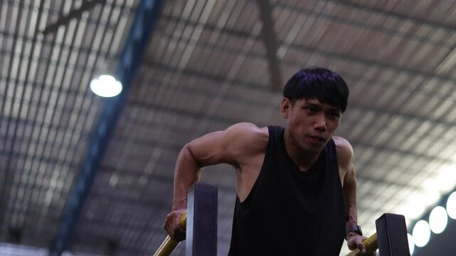 Muscular Asian man doing triceps dips on parallel bars at the gym.