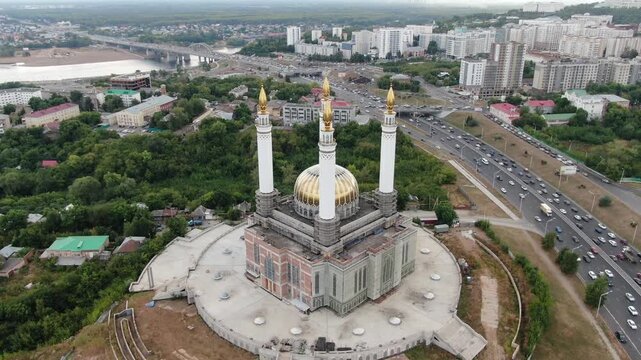 UFA, RUSSIA. AUG 12, 2022: Aerial view of the majestic Ar-Rahim Mosque with its golden dome and tall white minarets under construction, overlooking a busy highway and cityscape