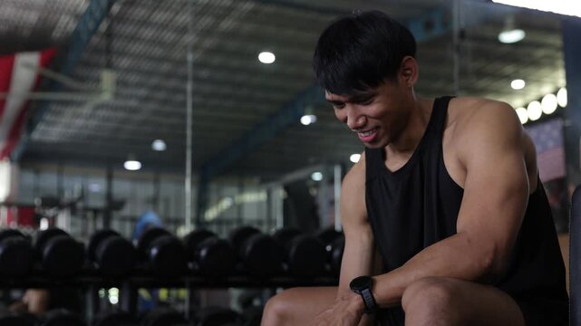Strong athletic Asian man in sportswear sitting on a bench and lifting a dumbbell for a bicep curl workout inside a fitness gym.