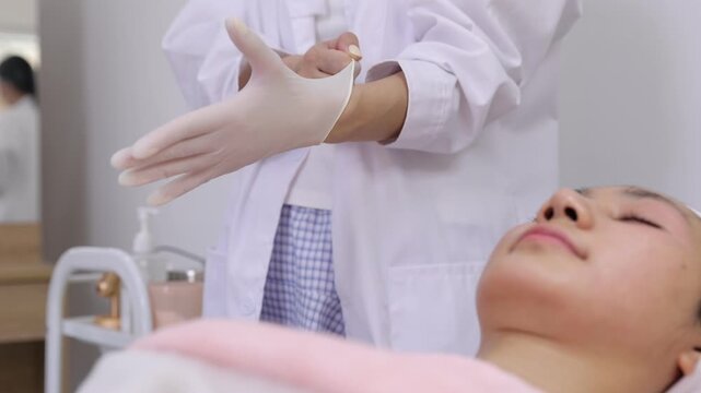 Esthetician putting on protective gloves before skincare procedure. Female beautician in a white coat putting on disposable latex gloves. A young woman client is lying on a bed in a beauty salon.