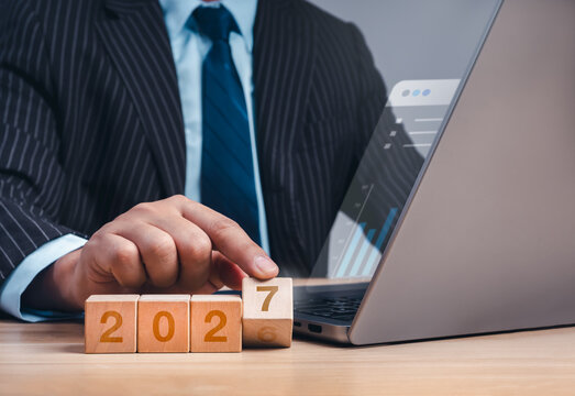 Business executive changes wooden number blocks from 2026 to 2027 beside laptop with performance graph, representing annual strategy, future planning, corporate goals, forecasting and business growth.