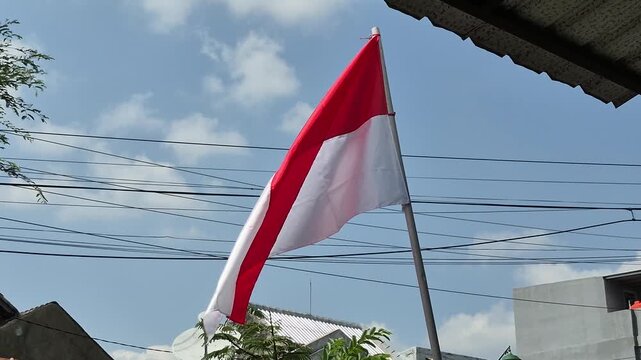 Red and white flag flutters against blue sky. Cable lines stretch across the frame. Dappled light catches the flag's fabric. Building rooftops frame the background