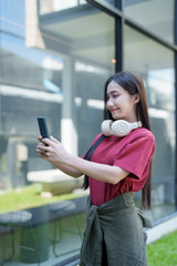 Fototapeta premium Young asian woman capturing a selfie with her smartphone while wearing headphones around her neck outdoors, reflecting modern communication, social media, and technology trends