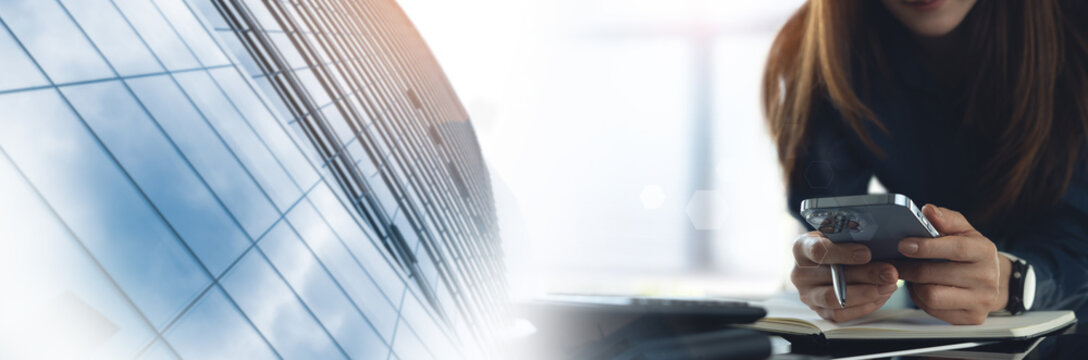 Business woman using smartphone with laptop and notebook on office desk with modern office building as background and copy space for business background