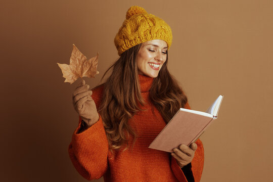 A woman in a yellow knit hat and orange sweater smiles peacefully with closed eyes, holding an autumn leaf and a book. This cozy image captures a joyful, serene fall moment.