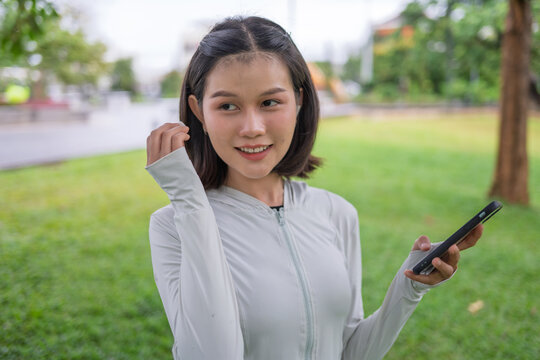Asian woman with wireless earbuds listening to music while warming up before jogging in urban park.