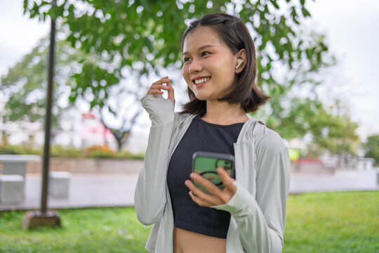 Asian woman with wireless earbuds listening to music while warming up before jogging in urban park.
