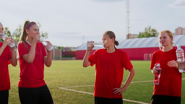 Young womens soccer team hydrating together during outdoor training session on green field, wearing matching red jerseys and resting between intense practice drills