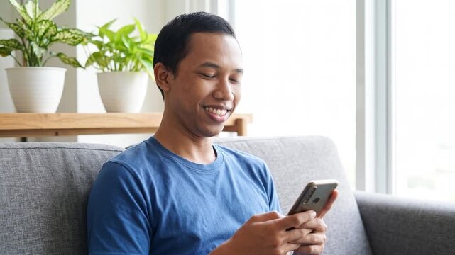Smiling happy young Indonesian man holding cell mobile phone using smartphone sitting at home on couch, scrolling social media, checking financial apps, buying online, texting messages. 