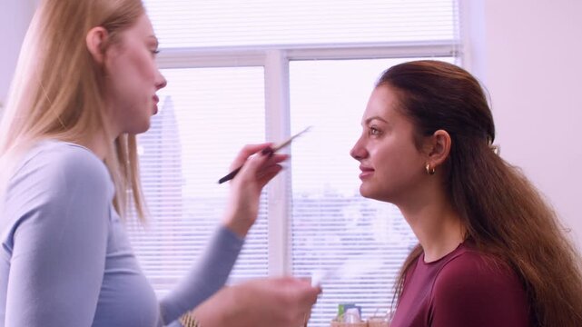 Face procedure. Cosmetic service. Makeup professional applying product cream for woman skin care in aesthetic studio indoor.