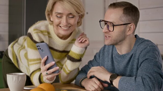 Middle aged Caucasian couple sitting at kitchen table, discussing content on smartphone and laughing. Perfect for mobile app marketing or digital lifestyle advertisements