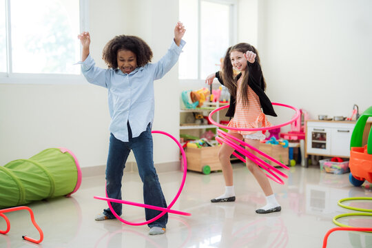 Energetic diverse young girls laughing and raising arms while playing with pink hula hoops in a kindergarten playroom, emphasizing active physical education, joy, and healthy child development.