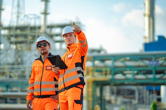 Industrial engineers discussing facility operations while pointing toward refinery equipment during site inspection. Engineering planning, safety management, asset monitoring, industrial maintenance.
