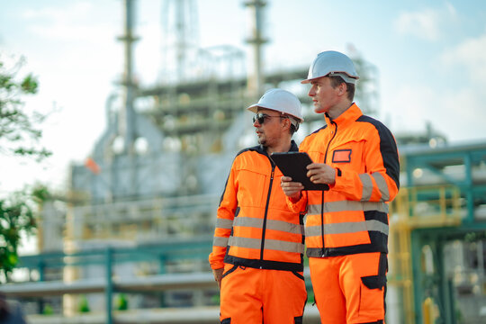 Two plant engineers holding a digital tablet while observing refinery infrastructure during an industrial inspection. Engineering teamwork, process monitoring, maintenance planning, workplace safety
