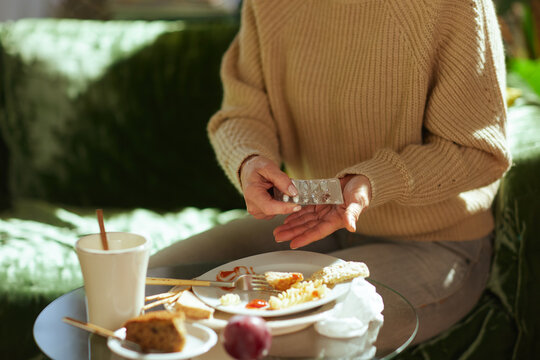 An adult woman holds a pack of pills over a table cluttered with takeaway leftovers and dessert, reflecting a routine of health management after an unhealthy meal.